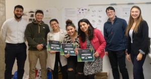 Participants of the Sustainable Building Challenge are standing in front of the project presentations. Three people in the center, Javeriya Hasan, Ike Arzoumanian and Eliany Rodriguez, are holding the certificates and smiling. 